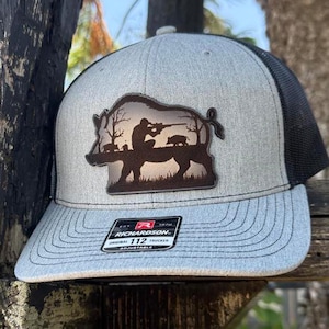Puede incluir: Gorra de camionero gris y negra con un parche que representa a un cazador apuntando con un rifle a un jabalí, con dos jabalíes más pequeños. La gorra tiene una visera curva y una parte trasera de malla. El parche dice "RICHARDSON 112".
