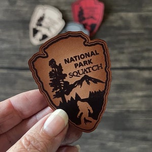 May include: Brown leather patch shaped like a National Park Service arrowhead with the text "National Park Squatch" and a silhouette of a Bigfoot walking in front of a mountain and tree.