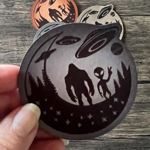 May include: A black and white round sticker featuring a silhouette of Bigfoot and an alien standing in a forest with a UFO in the sky. The sticker has a starry background.