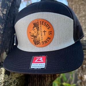 Puede incluir: Gorra snapback negra y beige con visera plana y parte trasera de malla. La gorra presenta un parche circular con un león coronado, una cruz y el texto "JESUS IS KING REVELATION 19:16". También tiene una etiqueta "FLAT BILL SNAPBACK".