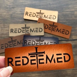 Peut inclure: Plusieurs patchs rectangulaires en cuir de différentes nuances de marron, beige et bleu. Chaque patch porte le mot "REDEEMED" en lettres noires grasses, avec un symbole de croix au-dessus du "E". Les patchs sont disposés sur une surface en bois.