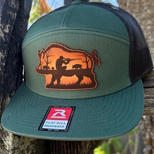May include: A green flat bill snapback hat with a black mesh back. The hat features a brown patch with a hunting scene of a hunter aiming at a boar. The patch is on the front of the hat. The hat has a tag that says "FLAT BILL SNAPBACK".