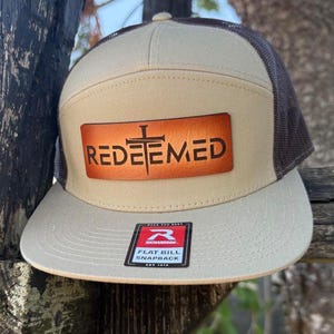 Puede incluir: Gorra snapback de visera plana marrón y beige con la palabra "REDEEMED" en letras marrones sobre un parche de cuero. La gorra tiene una parte trasera de malla y un parche Richardson.