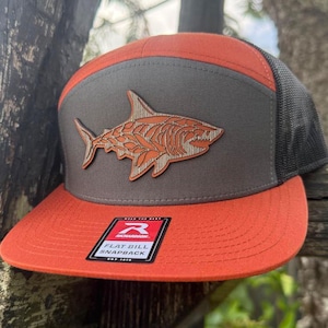 Puede incluir: Gorra snapback de visera plana naranja y gris con diseño de tiburón. La gorra presenta una parte trasera de malla y un parche con una ilustración de tiburón. La visera es naranja y el panel superior es gris. La gorra tiene el texto "FLAT BILL SNAPBACK".