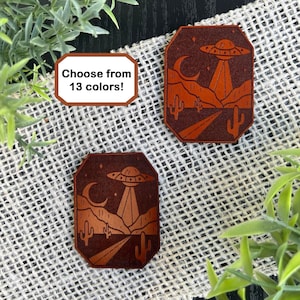 May include: Two brown leather coasters with a desert landscape and a UFO design. The coasters feature a crescent moon, stars, cacti, and a road. The text "Choose from 13 colors!" is on a white label.