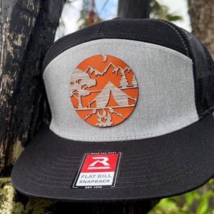 May include: Black and gray flat bill snapback hat with a mesh back. The hat features a circular wooden patch with a camping scene, including a tent, trees, mountains, and a campfire. The patch is orange and gray.