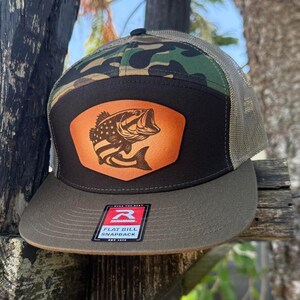 May include: A flat bill snapback hat with a camouflage top, a black front panel, and a tan mesh back. The front panel features a leather patch with a bass fish design. The hat has a curved brim and a snap closure.