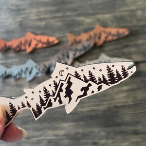 May include: A leather salmon silhouette with a mountain and forest design. The salmon is light brown with a black outline and the design is black. The salmon is on a wooden surface.