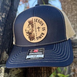 May include: Navy and beige trucker cap with a circular patch showing a lion, cross, and the words "JESUS IS KING". The cap has a curved peak and an adjustable strap. The brand is Richardson.