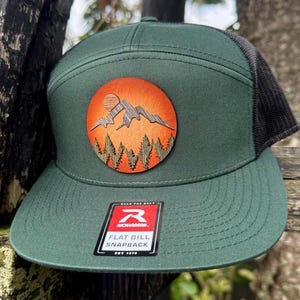 May include: Green flat bill snapback hat with a black mesh back. The hat features a round patch with a mountain and forest design in shades of orange and brown. The hat has a flat bill and a snapback closure.