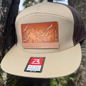 May include: A tan and brown flat bill snapback cap featuring a mountain and bear design on a leather patch. The cap has a mesh back and the text "FLAT BILL SNAPBACK".