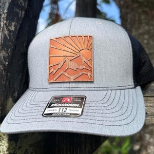 May include: Gray trucker hat with a black mesh back and a square, wooden patch featuring a mountain and sun design. The hat has a curved brim and an adjustable strap. The patch is made of wood and has a detailed mountain and sun design.