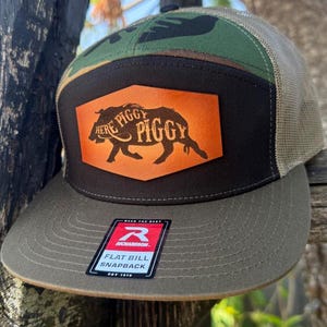 May include: A flat bill snapback hat with a camouflage top, a brown brim, and a tan mesh back. The front features a black patch with an orange background and a silhouette of a pig with the words "HERE PIGGY PIGGY".