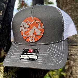 May include: Gray trucker hat with a white mesh back and a circular wooden patch featuring a camping scene with mountains, a tent, and a campfire. The hat has a curved brim and the text "RICHARDSON" on a tag.