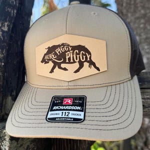 May include: A tan and black Richardson Original 112 trucker hat. The hat features a leather patch with a brown illustration of a pig and the words "Here Piggy Piggy". The hat is adjustable.