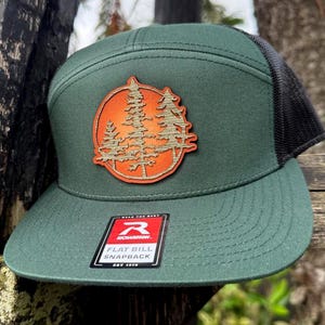 May include: Green flat bill snapback cap with a black mesh back. The cap features a circular patch with a forest scene in orange and brown. The cap has a Richardson tag that says "Flat Bill Snapback".