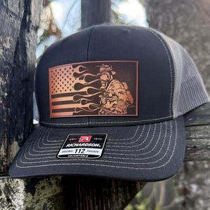 Puede incluir: Gorra de camionero negra y gris con un parche de cuero marrón que presenta una bandera estadounidense con llamas y un bombero. El parche dice "Richardson 112 Original Trucker Adjustable".