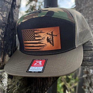 May include: A camouflage and black flat bill snapback hat with a brown leather patch featuring an American flag and a silhouette of a lineman working on a power line. The patch has the text "Rise to the Best". The hat has a mesh back and a green underside.