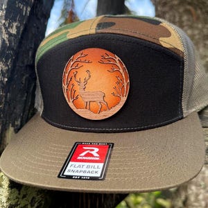Puede incluir: Gorra snapback de visera plana con parte superior de camuflaje, panel frontal negro y visera marrón. La gorra presenta un parche circular de madera con la silueta de un ciervo dentro de un diseño de árbol. La etiqueta dice "Wear the Best" y "EST 1970".