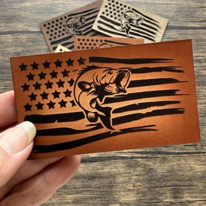 May include: A brown leather patch with a black silhouette of a bass fish jumping out of the water. The fish is superimposed on a distressed American flag design.
