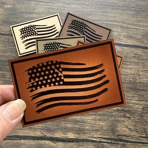 May include: A collection of rectangular leather patches featuring an American flag design. The patches vary in color, with the primary patch in a rich brown hue and black flag details. Other patches are beige and gray.