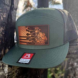 Puede incluir: Una gorra snapback de visera plana verde y negra con un parche de cuero que presenta a un bombero frente a una bandera estadounidense con llamas. El parche dice "Work Like the Best". La gorra tiene una etiqueta roja "Richardson" con el texto "Flat Bill Snapback".