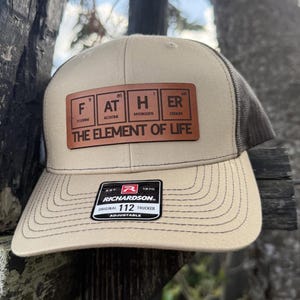 May include: A tan and black trucker hat with a brown leather patch that reads "FATHER - THE ELEMENT OF LIFE" with the letters representing the chemical elements of Fluorine, Astatine, Hydrogen, and Erbium.