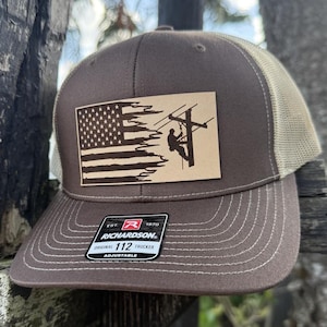 May include: A brown and tan trucker hat featuring an American flag and a silhouette of a lineman working on a power line. The hat has a leather patch with the text "EST. 1970 RICHARDSON. ORIGINAL TRUCKER 112 ADJUSTABLE".