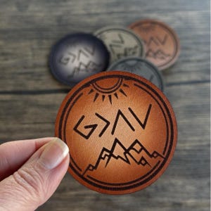 May include: A brown leather coaster with a sun and mountain design. The text "G>A" is in the center of the coaster.