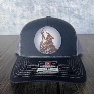 May include: A black and gray trucker hat with a circular patch featuring a howling wolf. The hat has a curved brim and a mesh back. The tag reads "RICHARDSON ORIGINAL 112 TRUCKER ADJUSTABLE."