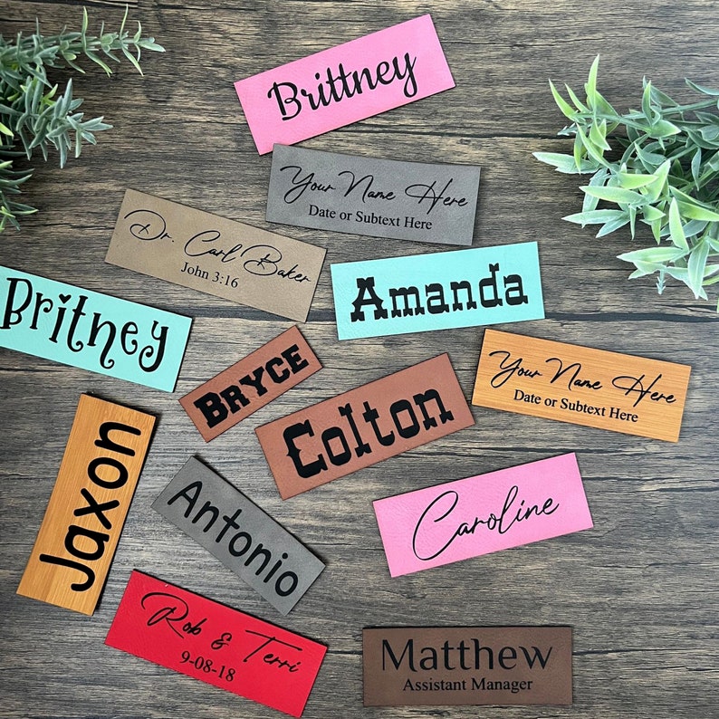 May include: Assortment of personalized leather name tags in various colors, including pink, brown, gray, red, and turquoise. The tags feature names like "Brittney", "Amanda", "Colton", "Antonio", "Matthew", "Caroline", "Bryce", "Jaxon", "Rob & Terri", and "Dr. Carl Baker". Some tags include additional text, such as "Your Name Here" and "Assistant Manager".