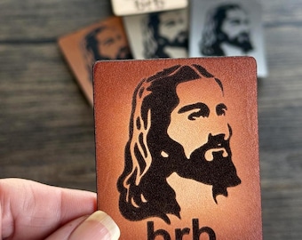 May include: A brown leather coaster with a black silhouette of Jesus and the text "brb" in white.