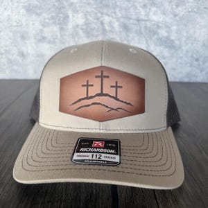 May include: A tan and black trucker hat with a brown leather patch featuring three crosses on a hill. The patch is a hexagon shape. The hat has a black mesh back and a brown adjustable strap with the text "Richardson" and "Original 112 Trucker Adjustable".