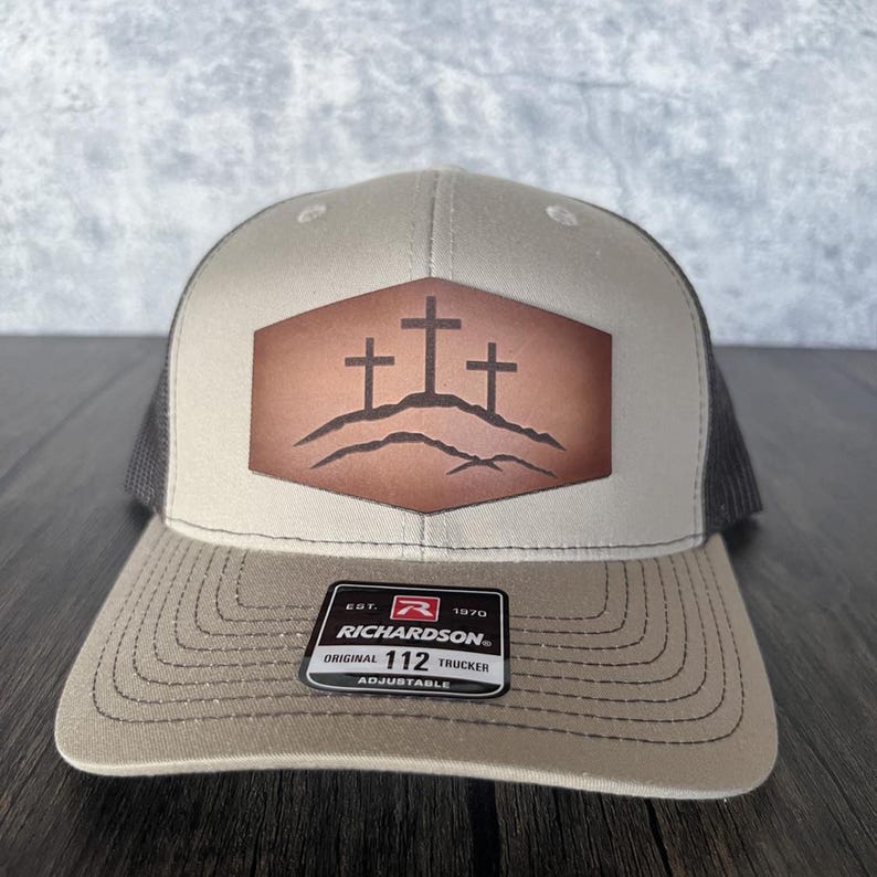 May include: A tan and black trucker hat with a brown leather patch featuring three crosses on a hill. The patch is a hexagon shape. The hat has a black mesh back and a brown adjustable strap with the text "Richardson" and "Original 112 Trucker Adjustable".