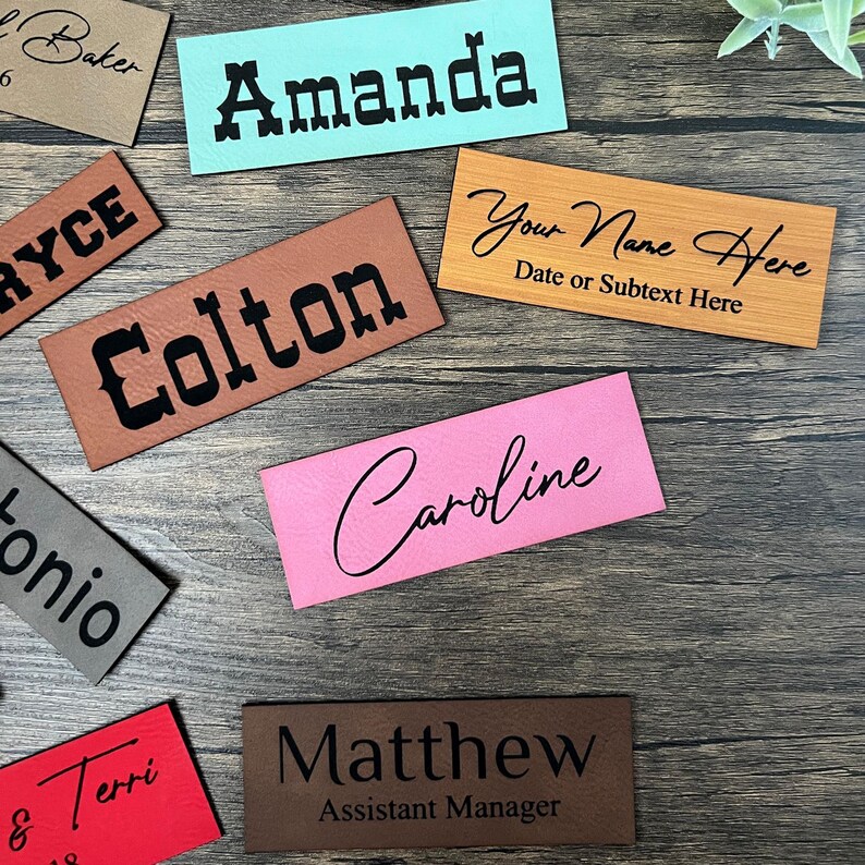 May include: Assortment of personalized leather name tags in various colors and fonts. The tags feature names like Amanda, Colton, Caroline, and Matthew. One tag has the text "Your Name Here" and "Date or Subtext Here".