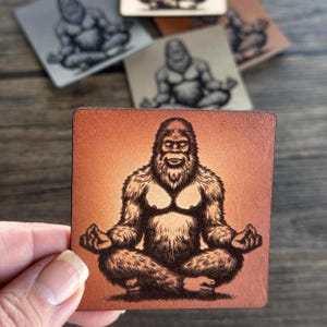 May include: A brown leather coaster with a black and white illustration of a Bigfoot sitting in a meditative pose with his hands on his knees.