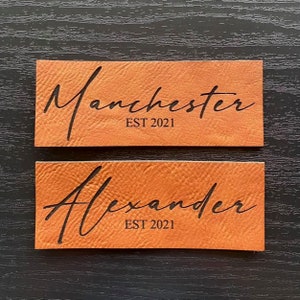Peut inclure: Deux patchs en cuir marron avec des lettres noires. Le patch du haut dit "Manchester EST 2021". Le patch du bas dit "Alexander EST 2021".