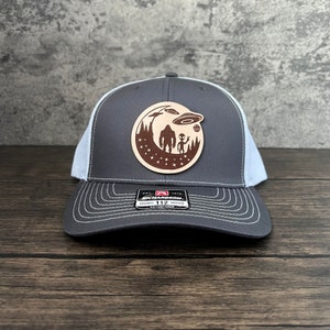 Peut inclure: Un chapeau de camionneur gris et blanc avec un patch en cuir marron représentant un Bigfoot et un extraterrestre debout devant un croissant de lune avec des étoiles et des soucoupes volantes. Le patch indique "Est. 1910 Richardson 112 Original 112 Trucker Adjustable".