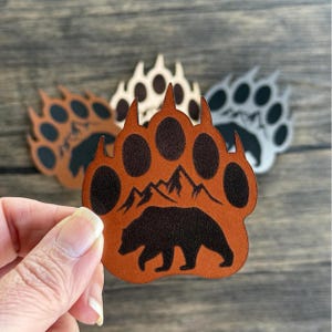 May include: Brown leather bear paw print patch with a silhouette of a bear and mountains inside the paw print.