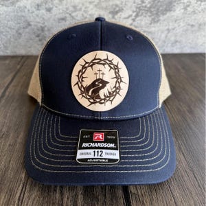 Peut inclure: Un chapeau de camionneur bleu marine et beige avec un patch en cuir brun représentant une couronne d'épines et deux croix. Le patch est centré sur le devant du chapeau. Le chapeau a une sangle réglable et le texte "EST. 1970 RICHARDSON. ORIGINAL 112 TRUCKER ADJUSTABLE" est imprimé sur une étiquette blanche attachée à la sangle.