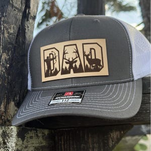 May include: A grey and white trucker hat with a brown leather patch that says "DAD" with a silhouette of a hunter with a bow and arrow and two deer. The patch is on the front of the hat.
