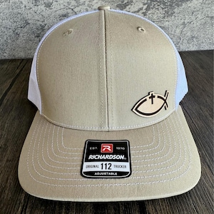 May include: A tan and white trucker hat with a brown leather fish symbol with a cross on it. The hat has a white mesh back and a black adjustable strap with the text "EST. 1970 RICHARDSON. ORIGINAL 112 TRUCKER ADJUSTABLE".