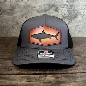 May include: A gray and black trucker hat with a brown leather patch featuring a shark silhouette. The patch has a hexagonal shape and the text "Richardson 112" is on the hat's underside.