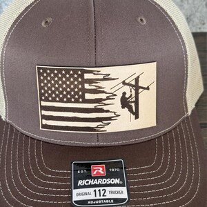 May include: Brown and beige trucker hat with a brown leather patch featuring an American flag with a distressed look and a silhouette of a lineman working on a power line. The patch has the text "EST. 1970 RICHARDSON" and "ORIGINAL 112 TRUCKER ADJUSTABLE".
