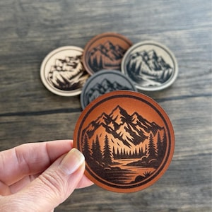 May include: Five leather coasters featuring a mountain range design. The coasters are brown, tan, and grey. The mountain range is black and the design includes trees and a river.