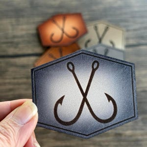 May include: A dark gray leather patch with a white gradient in the center. The patch features two crossed fishing hooks in black.