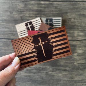May include: A brown leather patch with a distressed American flag design. A black cross with nails is centered on the flag. The patch is perfect for adding a patriotic touch to any project.