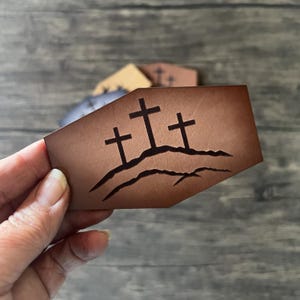 Peut inclure: Un objet en cuir marron avec trois croix noires sur un motif de colline. L'objet est tenu dans une main, avec d'autres objets similaires en arrière-plan. Le design est un symbole religieux simple mais puissant.