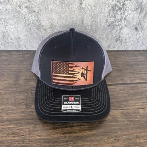 May include: A black and gray trucker hat with a brown leather patch featuring an American flag and a silhouette of a lineman working on a power line. The patch has the text "EST. 1970 RICHARDSON. ORIGINAL 112 TRUCKER ADJUSTABLE".