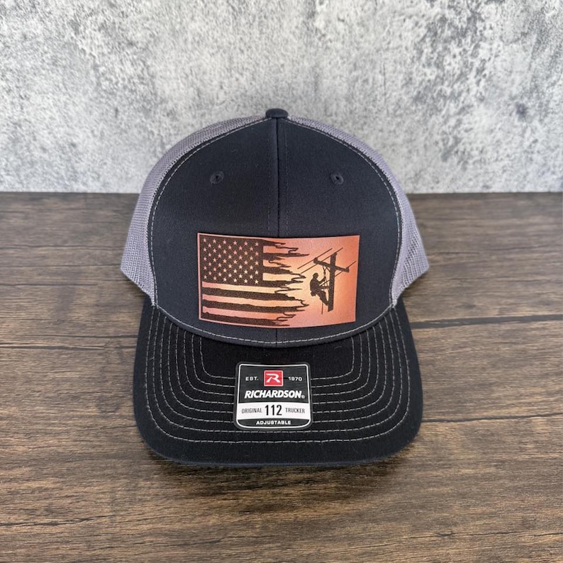 May include: A black and gray trucker hat with a brown leather patch featuring an American flag and a silhouette of a lineman working on a power line. The patch has the text "EST. 1970 RICHARDSON. ORIGINAL 112 TRUCKER ADJUSTABLE".